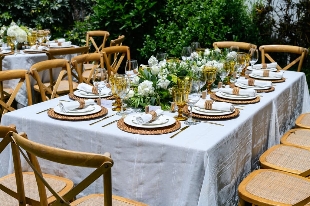 Elegant outdoor dining table set with white floral centerpieces, woven chargers, and wooden cross-back chairs.